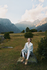 Fototapeta premium Young woman sits on a rock in a grassy meadow with distant mountains, savoring a peaceful outdoor moment, scene filled with soft light, warm tones and quiet contemplation