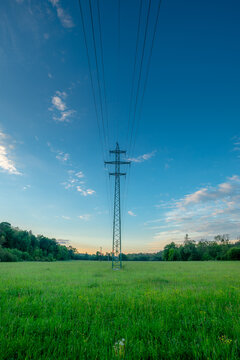 Tall steel electrical power transmission tower situated in a lush green countryside field, with thick overhead cables stretching across a bright blue sky during a peaceful summer day.