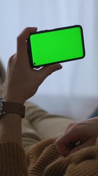 Person holding a smartphone with a green screen in a horizontal position, wearing a brown sweater and a wristwatch, set against a softly blurred indoor background