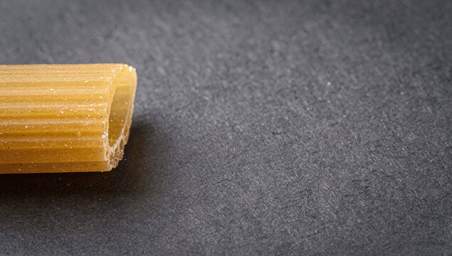 Single dry rigatoni pasta tube on a dark gray textured paper background with side placement