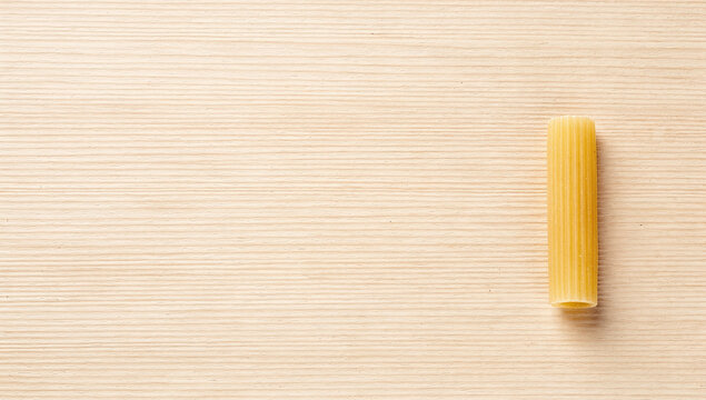 Single dry rigatoni pasta tube standing vertically on a light wooden table surface with side placement