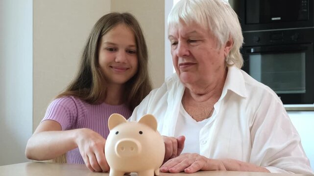 Young girl and grandmother putting money in piggy bank while sitting at kitchen table. Concept of wealth accumulation and bank services