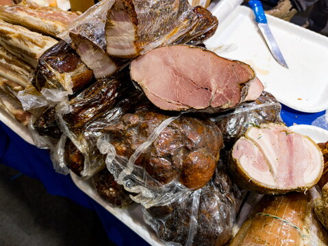 Selection of smoked and cured meats arranged on a market table with some pieces cut to reveal the texture and marbling inside. Vacuum-sealed packaging protects some meat portions, while others are unc