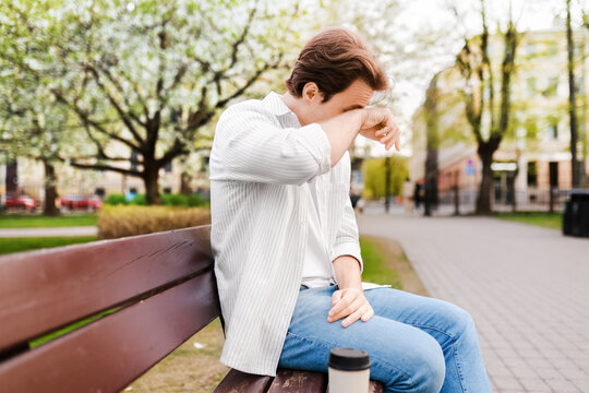 Man rubbing his itchy eyes while sitting outdoor in a spring park. Suffering from hayfever and a pollen allergy during bloom season.