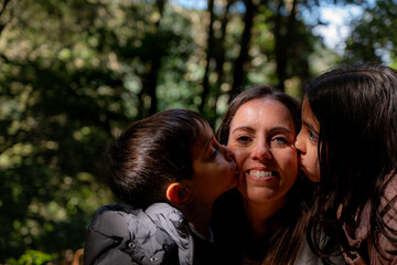 Obraz premium The mother smiles tenderly as her children kiss her on the cheeks. Sunlight filters through the trees behind them.