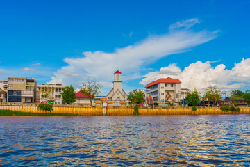 Obraz premium Riverside town with church tower, residential buildings, and blue sky with clouds reflecting on water surface in Chiang Mai Thailand