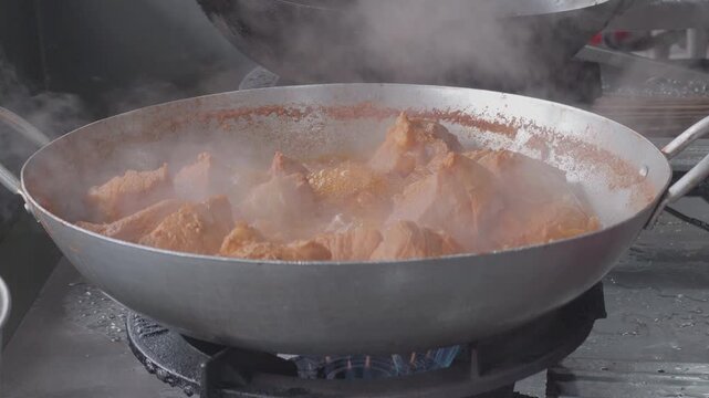Large chunks of seasoned pork meat simmering and boiling in a large wok on a gas stove with steam rising, close up of traditional street food cooking