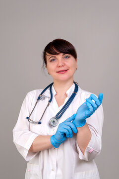Caucasian female healthcare professional wearing white coat puts on blue gloves, preparing for a medical procedure in a neutral studio setting with gray background