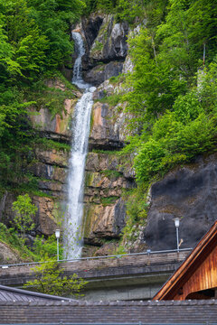 Tall alpine waterfall flowing over mossy rock face above village rooftops and lamps in springtime, Hallstatt, Austria.