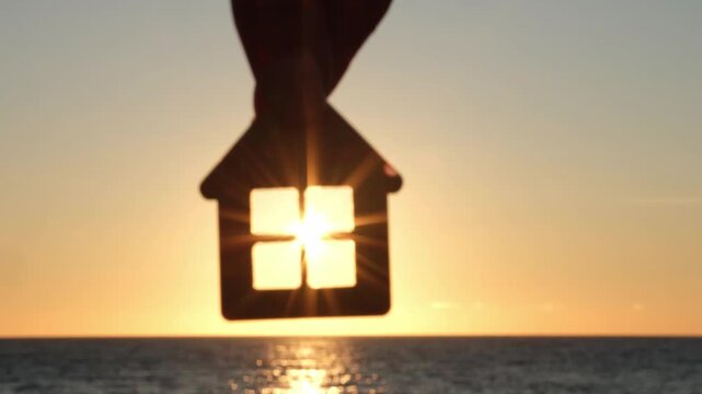 Hand holding house shaped keychain on background of sea sunset. Concept of mortgage loan, real estate purchase, property sale and rent