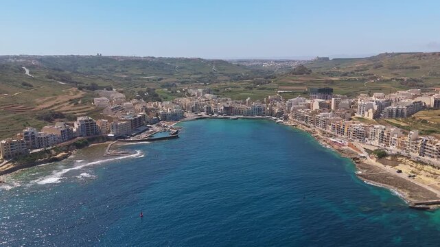 In Marsalforn on Gozo, fishing boats rest in a turquoise Mediterranean bay bordered by a waterfront of village buildings and rocky cliffs in a calm summer coastal setting.