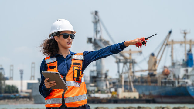 Woman wears a white hard hat and orange safety vest. She holds a tablet and points with a pen. She directs maritime freight operations at a commercial port. Cranes and ships appear in the background