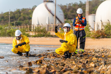 Fototapeta premium Three hazmat experts analyze toxic water pollution at a petrochemical refinery beach. One uses a tablet for data. Female ecologist observes the site. They wear protective suits and hard hats