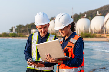 Two marine engineers in hard hats and safety vests review a laptop. They plan coastal environmental hazard control near oil storage tanks. ESG management for the energy sector