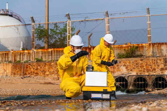 Two hazmat experts in yellow suits and white hard hats analyze toxic wastewater pollution with a laptop. They work near a large gas storage tank in a muddy industrial area