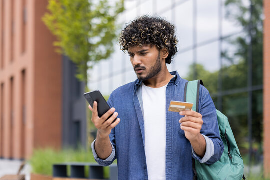Young man standing outdoors, frustrated and confused while trying to complete a mobile banking or online payment with smartphone and credit card, facing technical or data entry errors