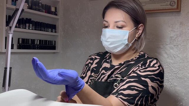 Woman wears mask and puts on blue gloves. She holds up one glove for display. She prepares for client treatment. Scene shows professional nail care setup. Her focus suggests calm, confident demeanor
