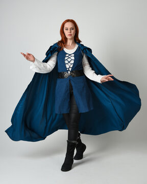 full length portrait redhead female model wearing medieval fantasy warrior historical costume with blue hood cloak. walking away with magical spell arm gesture, silhouette figure on studio background.