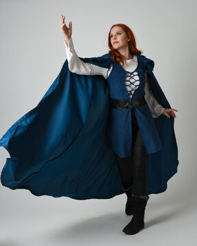 full length portrait redhead female model wearing medieval fantasy warrior historical costume with blue hood cloak. walking away with magical spell arm gesture, silhouette figure on studio background.