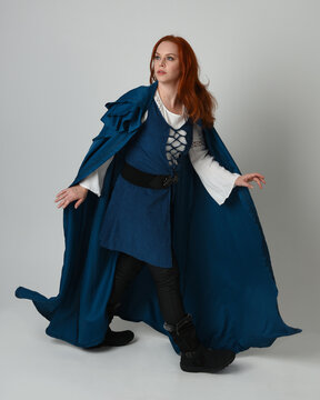 full length portrait redhead female model wearing medieval fantasy warrior historical costume with blue hood cloak. walking away with magical spell arm gesture, silhouette figure on studio background.
