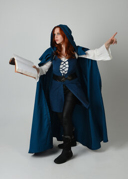 full length portrait of female model wearing medieval fantasy warrior historical costume with blue cloak. silhouette figure,  holding magical spell book on studio background.
