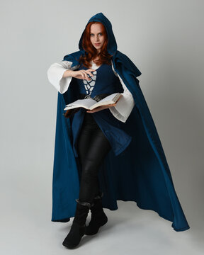 full length portrait of female model wearing medieval fantasy warrior historical costume with blue cloak. silhouette figure,  holding magical spell book on studio background.
