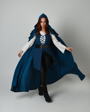 full length portrait redhead female model wearing medieval fantasy warrior historical costume with blue hood cloak. walking away with magical spell arm gesture, silhouette figure on studio background.