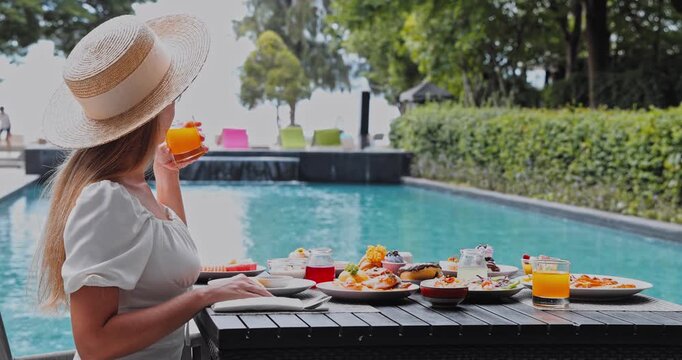 Woman wearing straw hat drinking orange juice at poolside breakfast table, tropical resort environment, luxury vacation and summer travel promotional content