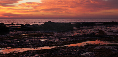 Crimson sky paints the ocean's edge with warm hues. Wet rocks glisten, mirroring the upcoming daylight. Sunrise casts a soft glow across the horizon's edge © WendlerCanning Photo