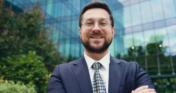 Close-up of a confident businessman in a formal suit with crossed arms near a glass office building, looking at the camera and smiling outdoors.