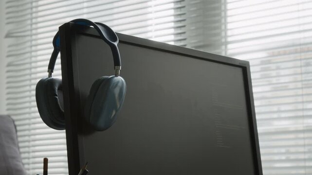 No people medium pan shot of wireless headphones hanging off computer screen displaying lines of code on black background during workday, white window blinds in background,