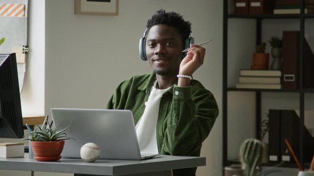 Medium portrait of young African American male software engineer focusing on work in startup office on laptop, listening to music in headphones, looking at camera, smiling while fiddling with pen