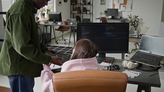 Medium rear shot of junior African American male web developer walking up and showing code on tablet, while female Caucasian supervisor checking and circling components for correction and improvement