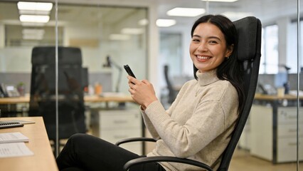 Fototapeta premium Smiling businesswoman relaxes in office chair while checking smartphone during workday.