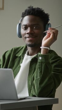 Medium vertical portrait of African American male web developer in headphones working on computer in office, concentrating on project task while fiddling with pen, looking up at camera, smiling