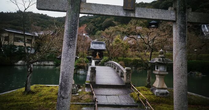 Tall stone torii gate near small bridge in Japanese village as it rains - steady cam tracking shot