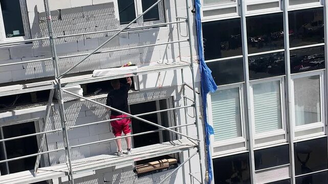 Construction worker on scaffolding installing windows on a modern building facade. A person in red shorts works on a building exterior, surrounded by scaffolding and new window frames, indicating