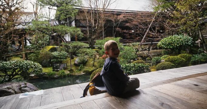 Happy little boy sitting on porch of ancient Japanese home with beautiful garden ahead - steady cam shot