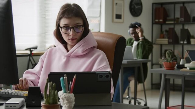 Medium shot of young Caucasian female software developer writing code on desktop computer while working in corporate office, testing execution on tablet, black male colleague working in background