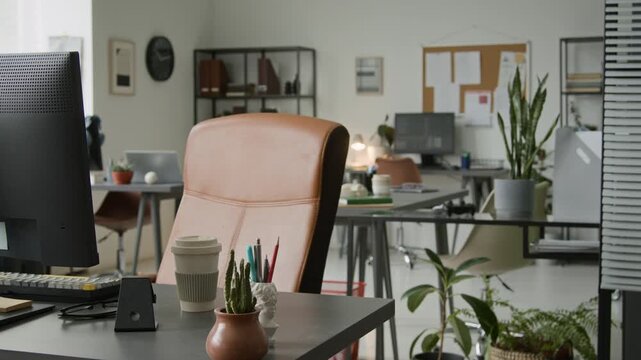 Medium pan no people shot of workplace of programmer in startup company office, comprising desk, computer with large screen, comfortable leather chair, coffee cup, green potted plants, before workday