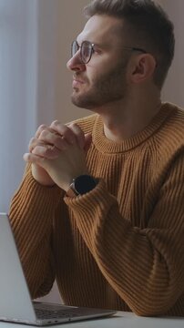 Young man wearing glasses and a brown knitted sweater sitting thoughtfully in front of a laptop, engaged in deep thinking or focused work by a window with soft natural light