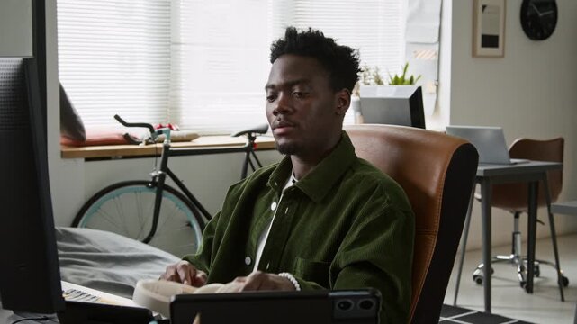 Medium shot of young African American web developer putting on headphones, turning on music on smartphone for inspiration, smiling and relaxing in chair while working on computer code for new project