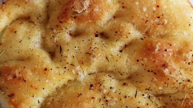 Macro shot panning across the golden crust of freshly baked Italian focaccia bread topped with herbs, salt, and spices. Detailed view of the textured surface showing rosemary, pepper, and seasonings o