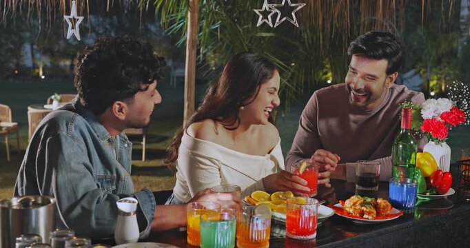 Indian young adult gen z relax male female sitting around bar table talk speak have fun joy trip happy girl boy man pick up glass do cheers drink juice spend good time give high five enjoy night place