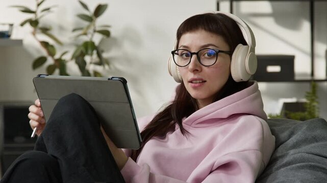 Medium close-up portrait of young Caucasian zoomer female developer working on tablet computer while designing website or mobile app, chilling out in bean bag chair, looking at camera with smile