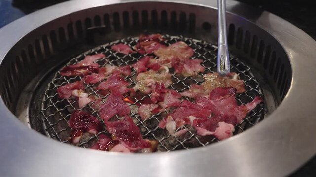 Thin slices of raw beef grilling on a traditional round wire mesh charcoal barbecue. Metal tongs handle the meat over hot coals. Authentic Japanese yakiniku or Korean BBQ dining experience.