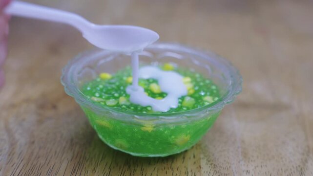 Traditional Thai dessert sago with corn and coconut milk being poured from a spoon into a glass bowl on wooden background. Sweet tapioca pearls with coconut cream topping and jelly texture.