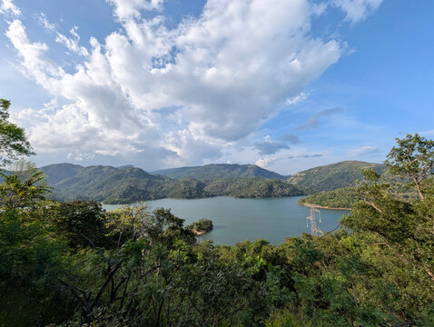 Panoramic vista of a large freshwater reservoir winding through green tropical mountain ridges under bright, heavy clouds.