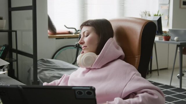 Medium shot of sleepy Caucasian female programmer stretching, sitting up in chair, putting on eyeglasses while writing code for mobile application on computer, laptop and tablet on desk