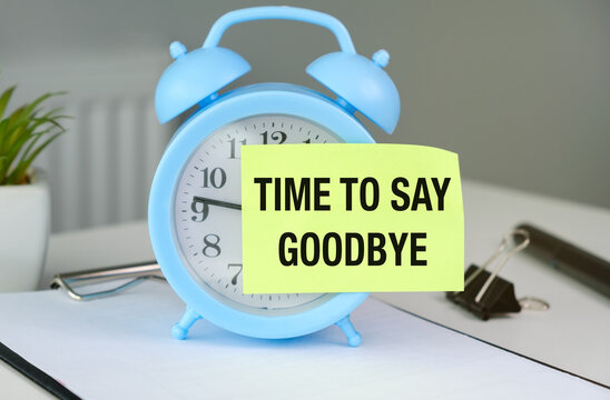 Blue alarm clock sits on a desk. Yellow sticky note reads TIME TO SAY GOODBYE. White notebook lies beside the clock.